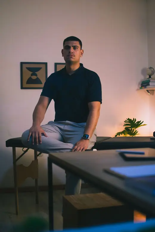 Felipe sentado na maca do consultório, com a mesa de trabalho à frente